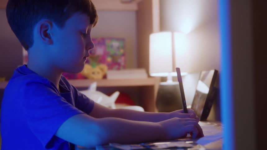 The video is a medium closeup of a young boy with dark hair and a blue tshirt looking intently down at a device or book he is seated in a dimly lit room with his face illuminated by a bright cooltoned light source from the screen