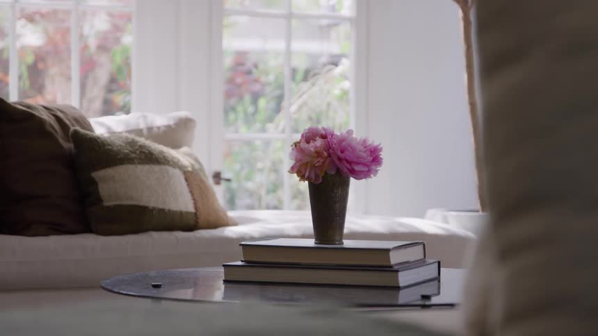 A still medium view of a glass coffee table features a vase of pink peonies on two stacked books set against a bright window and a white sofa with neutral pillows creating a cozy and peaceful domestic atmosphere