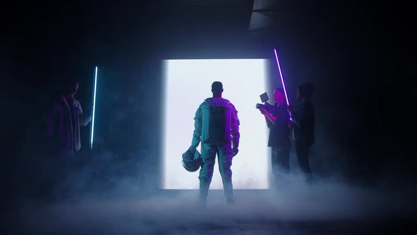 The video is a wide shot of an astronaut in a white space suit standing in a dark foggy room they are facing a bright rectangular portal of light while other figures holding neoncolored light swords stand around the edges