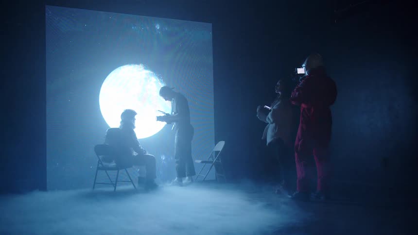 Four people stand in a dark hazy room with a moon image projected onto a rear wall while one person sits and another stands near the screen and the remaining two people stand to the right with one holding a recording device