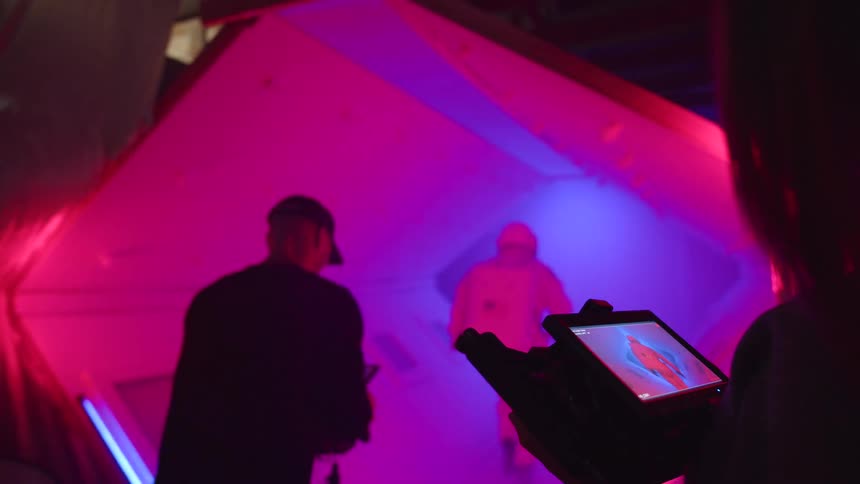The video shows a person in a dark silhouette standing in front of a large angular object bathed in bright red and magenta neon light the person is holding a small tablet or screen which is also glowing with bright colorful light
