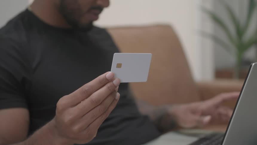 The video is a closeup shot of a mans hands holding a white debit or credit card he is seated wearing a black tshirt and appears to be in the process of entering information into a laptop visible on his lap