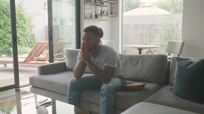 Man sitting in his living room with popcorn sits up and covers his face upset