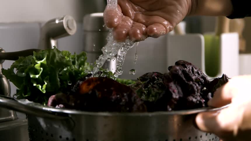 A persons hand is positioned under a stream of water flowing from a metal faucet into a metal strainer holding dark red root vegetables and green leafy plants