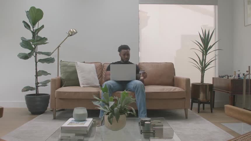 The video shows a man sitting on a tan couch in a modern welllit living room and working on a laptop the room features a glass coffee table with a plant a tall floor lamp and other large houseplants