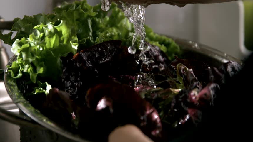 The video is a closeup shot of hands rinsing a large metal bowl filled with fresh leafy greens and red root vegetables under running water the water is pouring from a faucet and cascades over the produce against a dark background