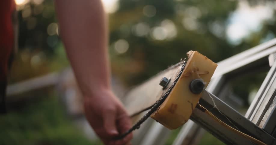 A persons hands manipulate a metal chain around a bolted guide piece on a larger metal object with the work taking place outdoors against a blurred background