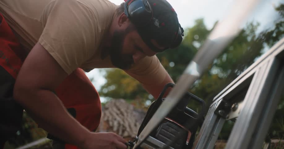 A man wearing ear protection and an orange safety vest kneels over a power saw mounted on a metal support frame working to saw a piece of wood while positioned outdoors against a background of green trees