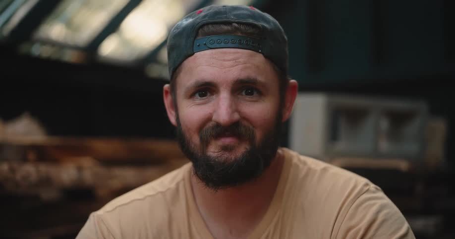 The video is a medium closeup of a bearded man wearing a tan tshirt and a dark baseball cap with red accents he is first looking down and then looks up directly at the camera maintaining a serious expression