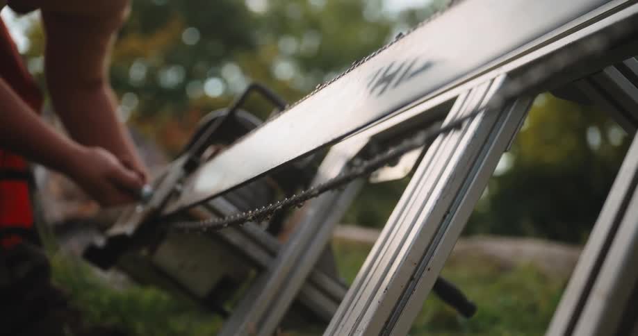 A person in an outdoor location uses their hands and a small tool to adjust the placement of a toothed metal chain around a long flat metal guide bar of a power saw which rests on a metal stand