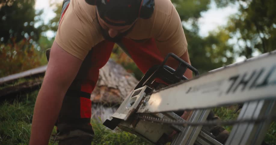 The video is a closeup of a person with gloves and an orange work suit adjusting the chain and guide bar of a chainsaw resting on a metal frame the scene is outdoors with green grass and soft natural lighting