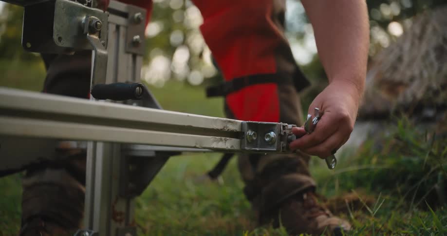 The video is a mediumclose shot of a man wearing a tan tshirt orange pants and ear defenders over a baseball cap he is bending down to adjust a piece of logging equipment that is holding a chainsaw