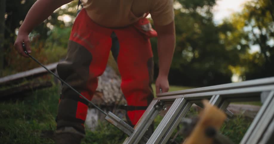 A worker wearing a tan shirt orange protective chainsaw chaps and earmuffs bends over to work on the metal frame of a sawmill outdoors on green grass under bright lateafternoon sunlight