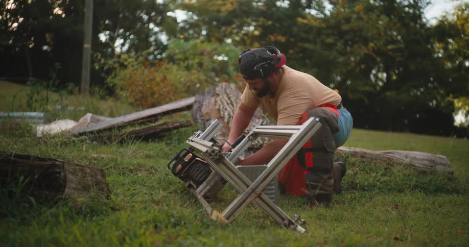 A man wearing safety ear protection and work pants kneels on a grassy field and leans forward to adjust a power saw that is mounted within a low silver metal framework with tree stumps and cut wood pieces lying nearby