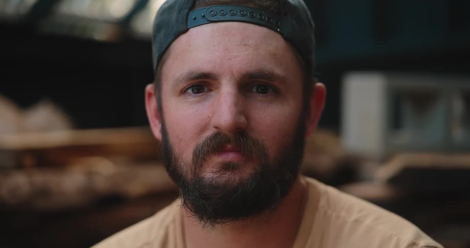 Man wearing backward cap looks on in setting with stacked wooden logs