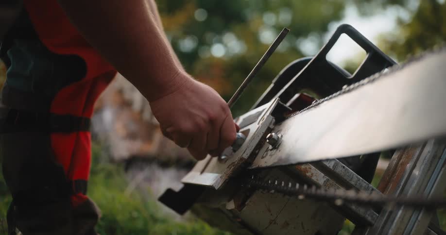 The video is a closeup of a person sharpening a chainsaw blade that is mounted on a sawhorse or guide rail the person is wearing orange highvisibility pants while working on the chain