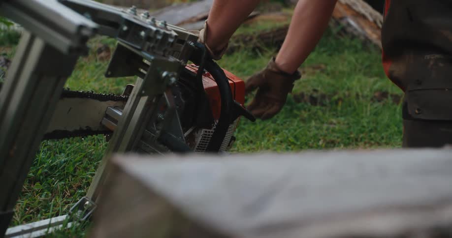 A close view of a worker wearing gloves and protective pants repeatedly pulls the cord to start a large gasoline chainsaw mounted to a guide frame outdoors on the grass