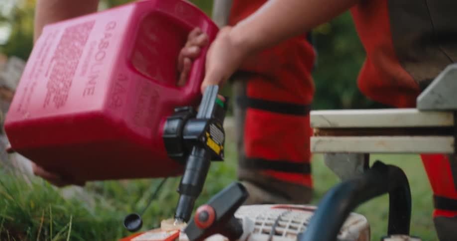 A bearded man wearing a tan shirt orange protective gear and earmuffs pours gasoline from a large red plastic can into the tank of an orange chainsaw with the operation taking place outdoors on the grass