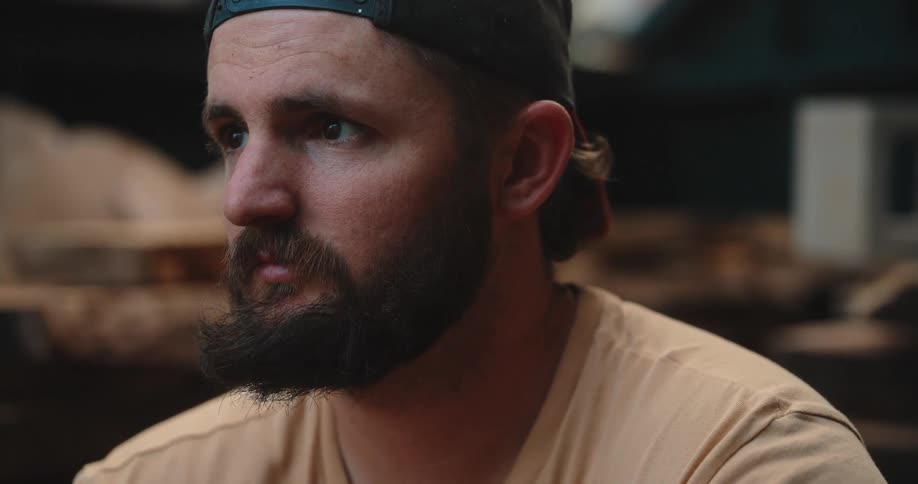 The video is a medium closeup shot of a man with a thick beard and dark baseball cap looking offcamera with a contemplative expression he is wearing a tan shirt and the background appears to be an indoor woodworking or shop setting
