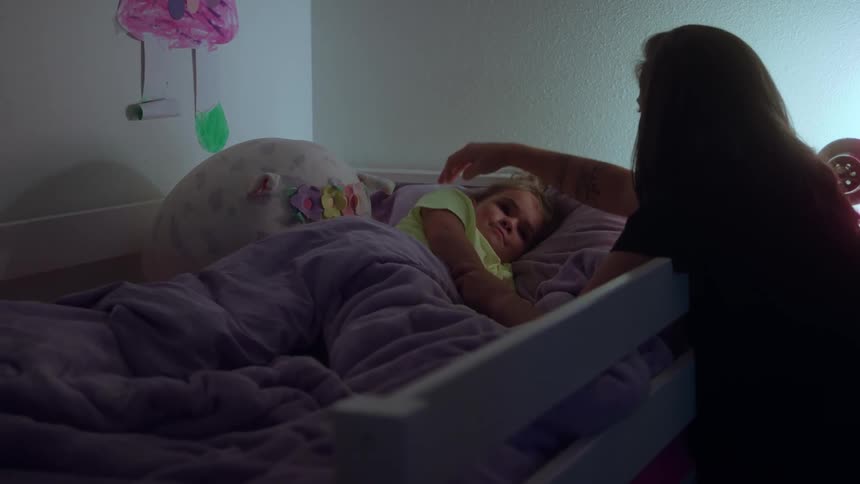 The video is a dimly lit interior shot of a mother standing next to a child tucked into a bunk bed the mother is gently stroking the childs head as they lie under a purple blanket