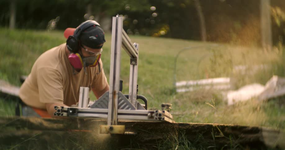 The video shows a man outdoors wearing headphones and a respirator mask operating a power tool to saw a wooden plank sawdust and wood chips are being kicked up by the saw as he works suggesting a lumber milling or processing activity