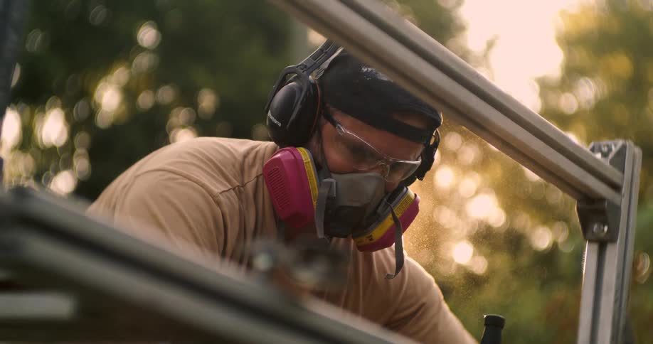 A carpenter wearing a respirator with magenta filters a black cap and black earmuffs bends over the metal frame of a portable sawmill as fine sawdust and bright sunlight filter through the air outdoors