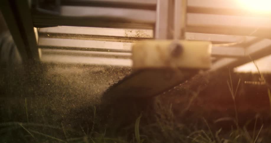 The video is an extreme closeup shot of a chainsaw blade cutting through a log positioned on a portable aluminum guide sawdust and wood chips are kicking up violently toward the camera and into the surrounding grass with bright sunlight flaring