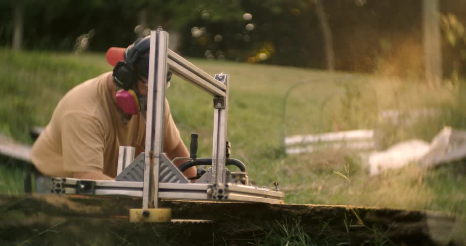 The video shows a man working outdoors in a grassy area using a saw mounted on an aluminum guide rail to cut a large piece of wood he is wearing a tan shirt protective earmuffs and a dust mask as he operates the equipment