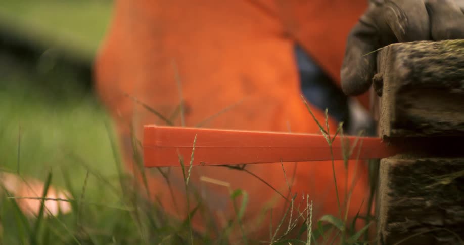 A close view of a worker wearing gloves and orange protective gear uses a red rubber mallet to gently hammer an orange plastic felling wedge into a log outdoors on the ground in the green grass