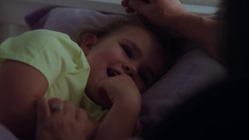 A close dark view shows a young girl lying in bed wearing a pale green shirt as a womans hand gently strokes the girls forehead creating an atmosphere of comfort and care