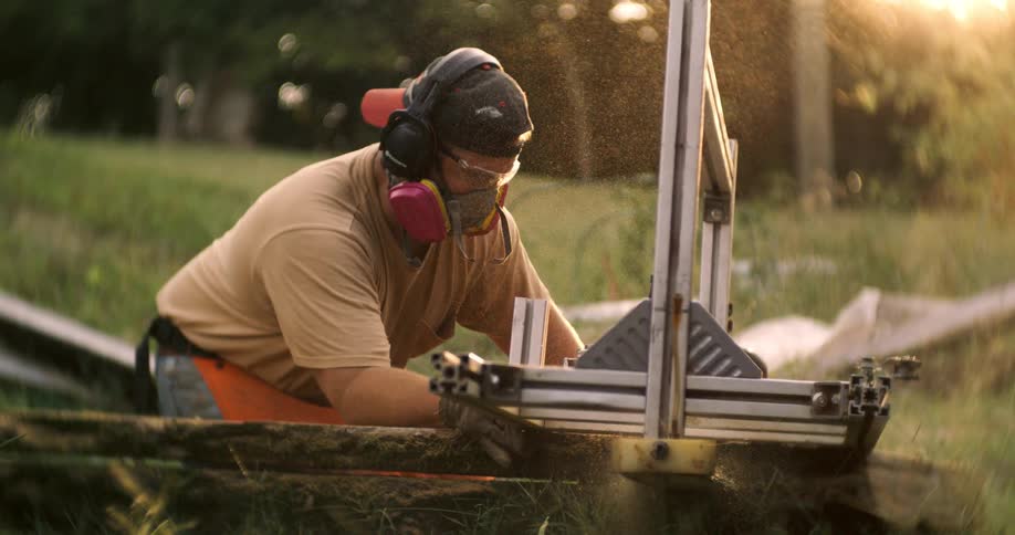 A man wearing full protective gear operates a chainsaw mill surrounded by a cloud of glowing sawdust