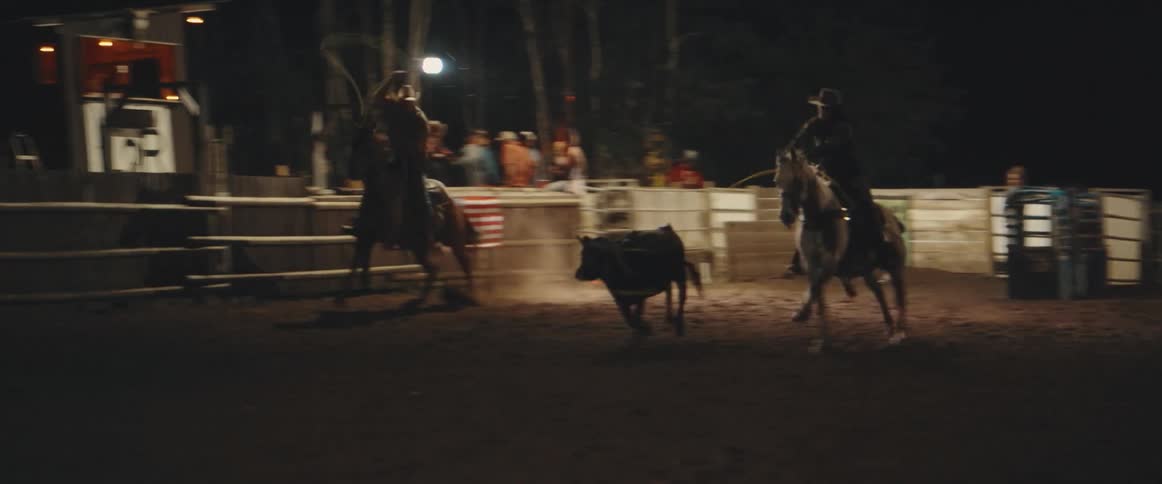 Riders on horseback rope steer in nighttime rodeo arena with audience in the background