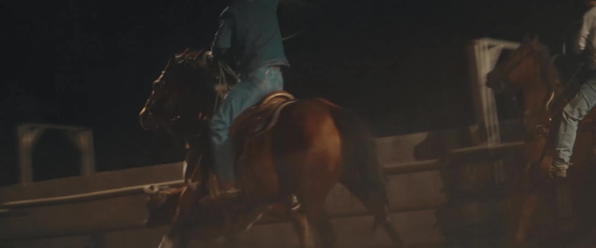 Two people ride horses on a dirt surface within a fenced enclosure with one rider swinging a rope overhead and another rider guiding a young cow in the foreground all taking place at night under artificial lighting