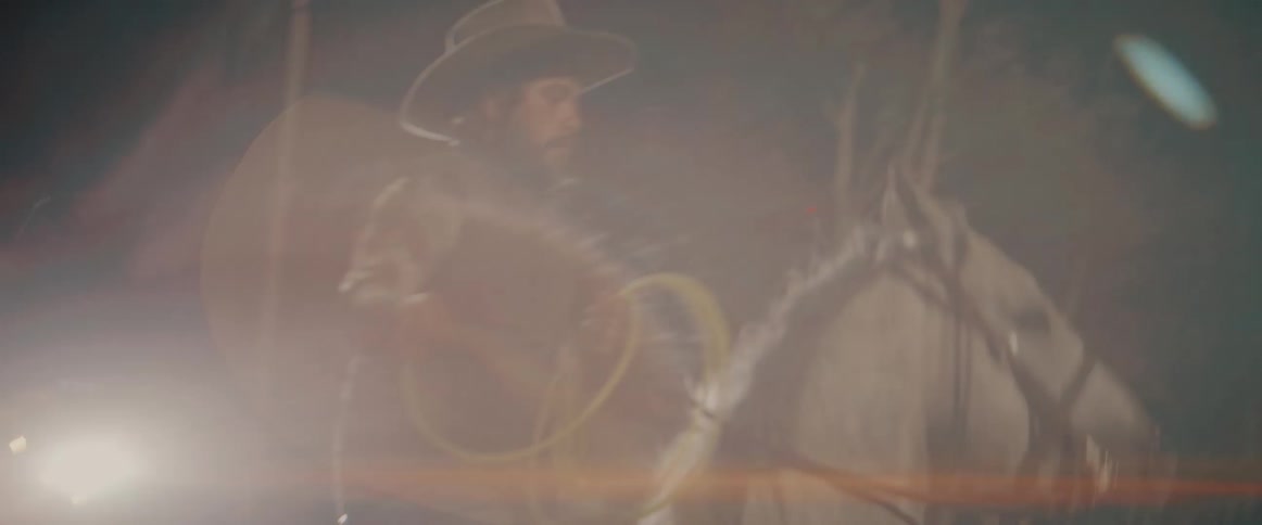 A person wearing a widebrimmed hat rides a lightcolored horse in a dark fenced enclosure while holding and swinging a coil of yellow rope with bright artificial light
