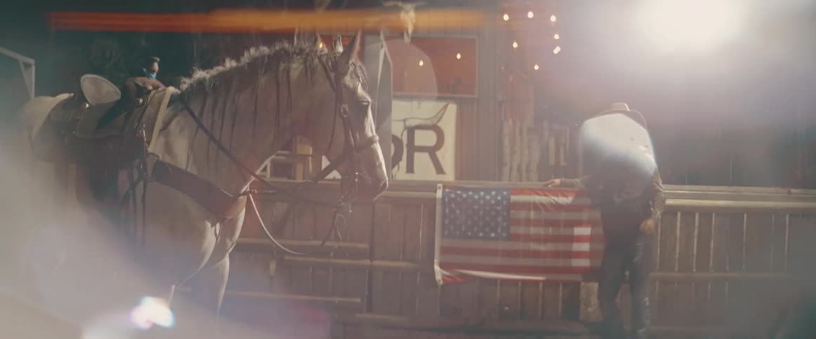 A cowboy wearing a hat and vest dismounts a lightcolored horse positioned near a wooden fence while a folded american flag hangs on the fence under artificial nighttime lighting