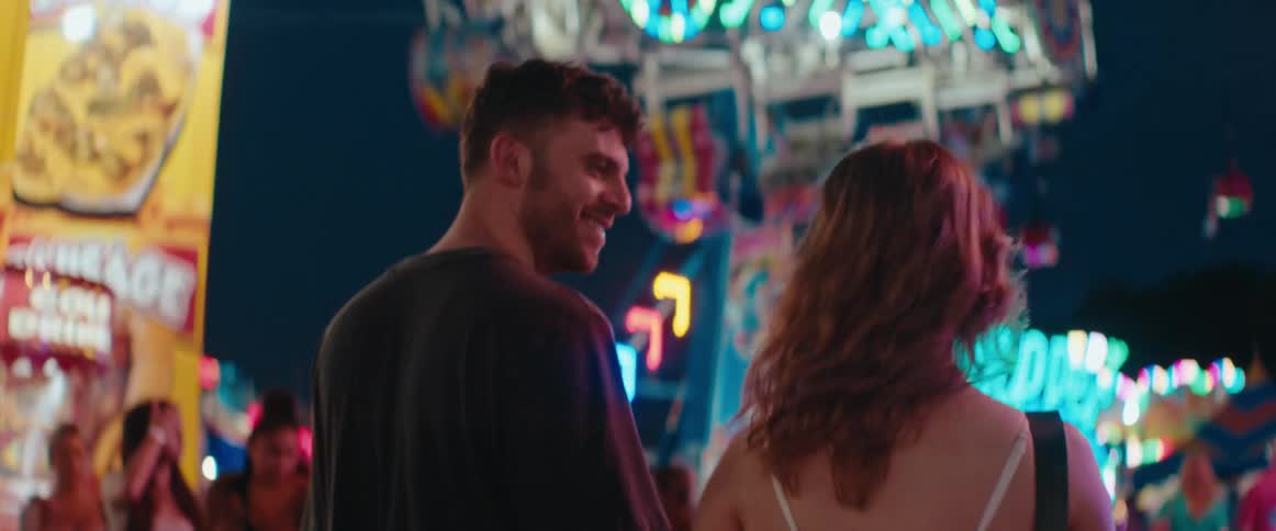 A man in a dark shirt and a woman in a light top walk sidebyside with their backs mostly to the camera looking at the illuminated structures of an amusement park in the background at night