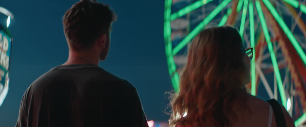 A man and a woman walk through a night carnival with neon lights and a ferris wheel glowing behind them