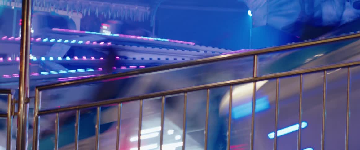Intense blue and pink neon lights from a fast spinning carnival ride blur into motion streaks behind a metal safety railing at night