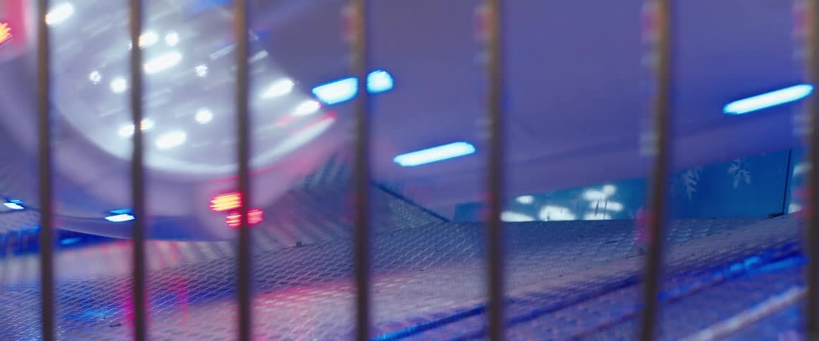A low angle view through metal bars shows the spinning platform of a carnival ride covered in a diamond plate pattern illuminated by bright neon lights that blur with the motion