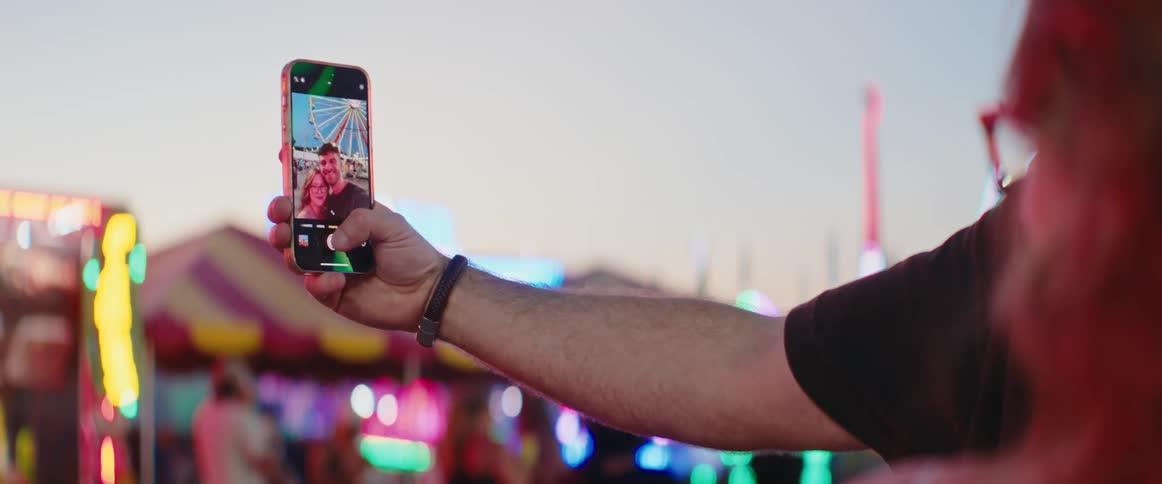 A person holds up a smartphone to take a selfie video of a man and a woman at a bright colorful fair or carnival with a ferris wheel visible in the distance during the twilight hour