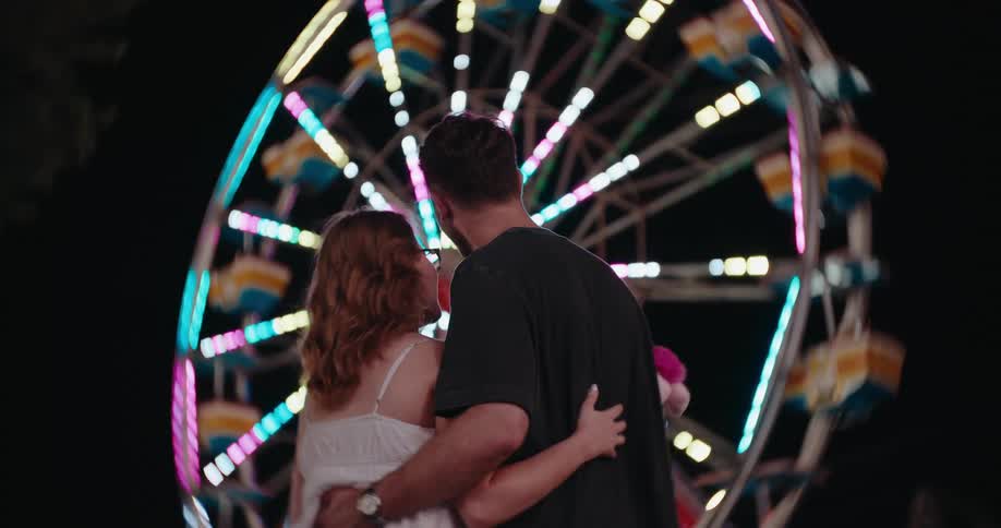 A couple embraces while watching a colorful illuminated ferris wheel at a carnival at night