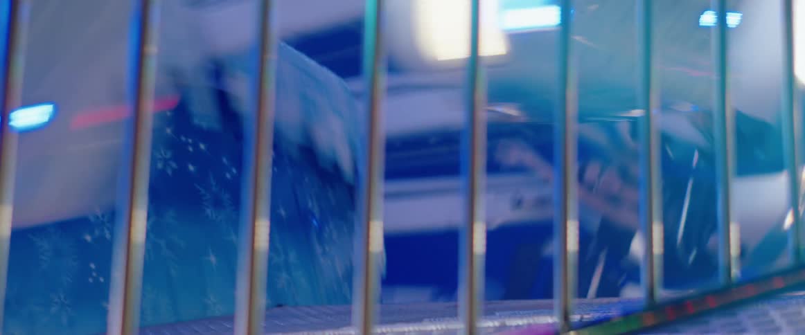 A spinning amusement ride flashes blue and white lights behind metal bars as reflections warp across its glossy surface
