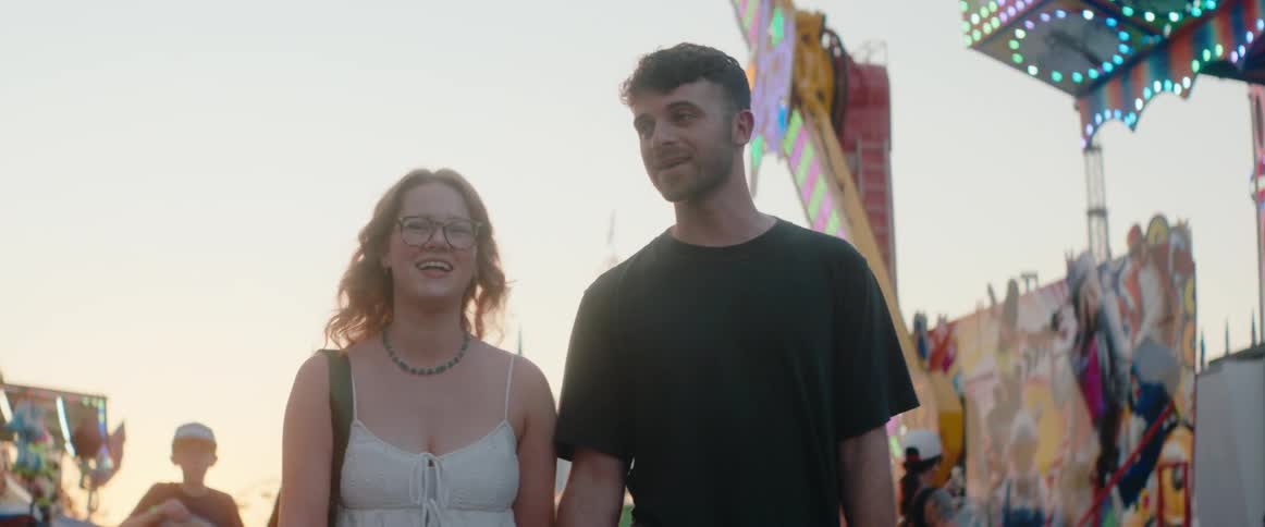 A man and a woman walk sidebyside in an outdoor location speaking and smiling while the background shows the moving illuminated structures of several amusement park rides