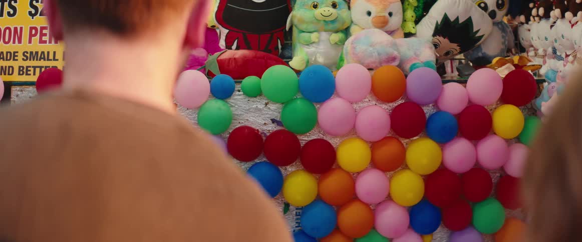 A persons shoulder and arm are visible in the foreground as they move to throw an object at a densely packed wall composed of brightly colored oval balloons with rows of large stuffed animal prizes displayed directly above the target area
