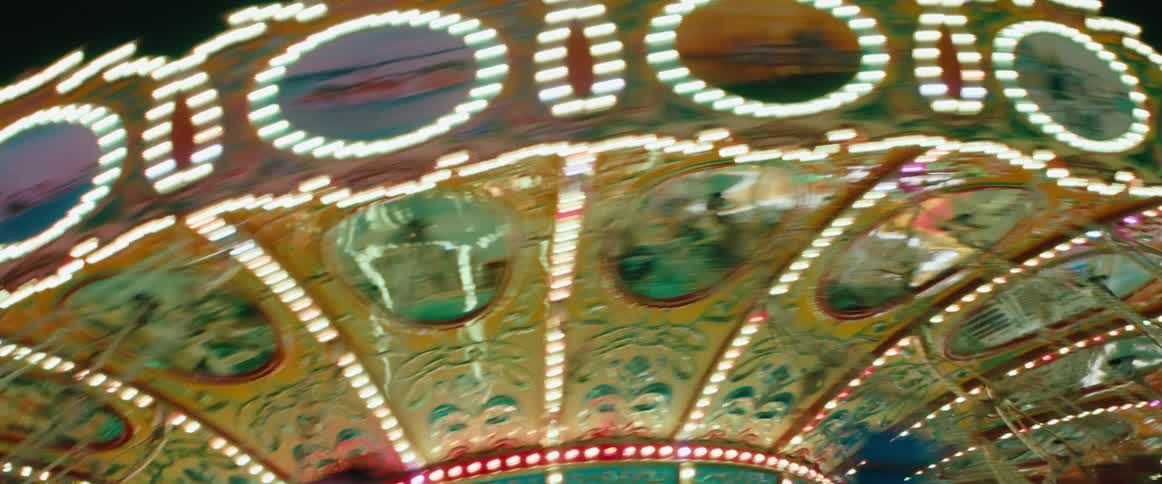 A brightly lit carnival swing ride whirls at night its ornate painted panels and strings of bulbs streaking into colorful blur