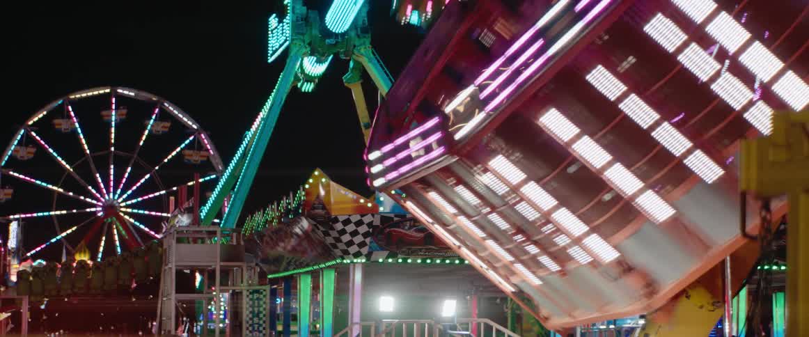 A camera is focused on a vertically swinging boxshaped amusement park ride lit by rapidly changing colored lights in the foreground with a large rotating wheel ride and a tall swinging pendulum ride in the background all operating at night