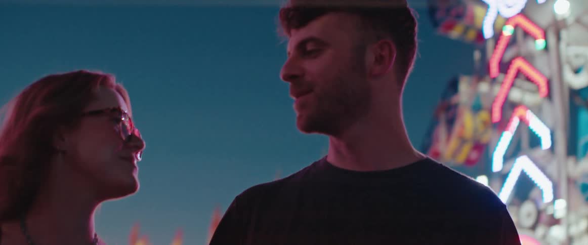 At a carnival at night a man affectionately kisses a smiling woman on the forehead