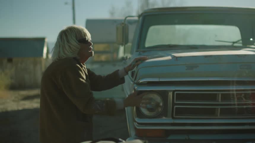An elderly woman rubs her hands on an old pickup truck
