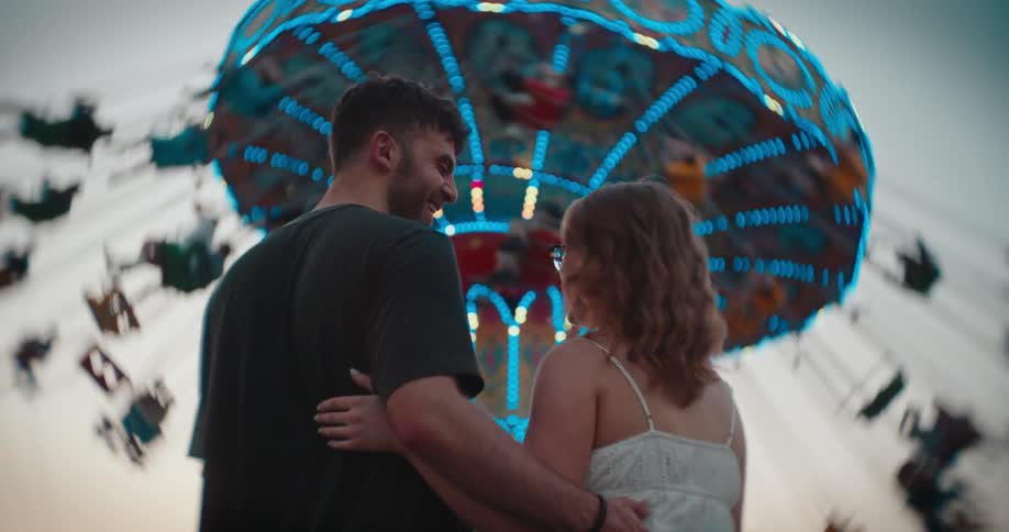 A man and a woman stand close to each other with their arms around one anothers waist and back facing each other and smiling while a large brightly lit swing ride rotates rapidly in the immediate background