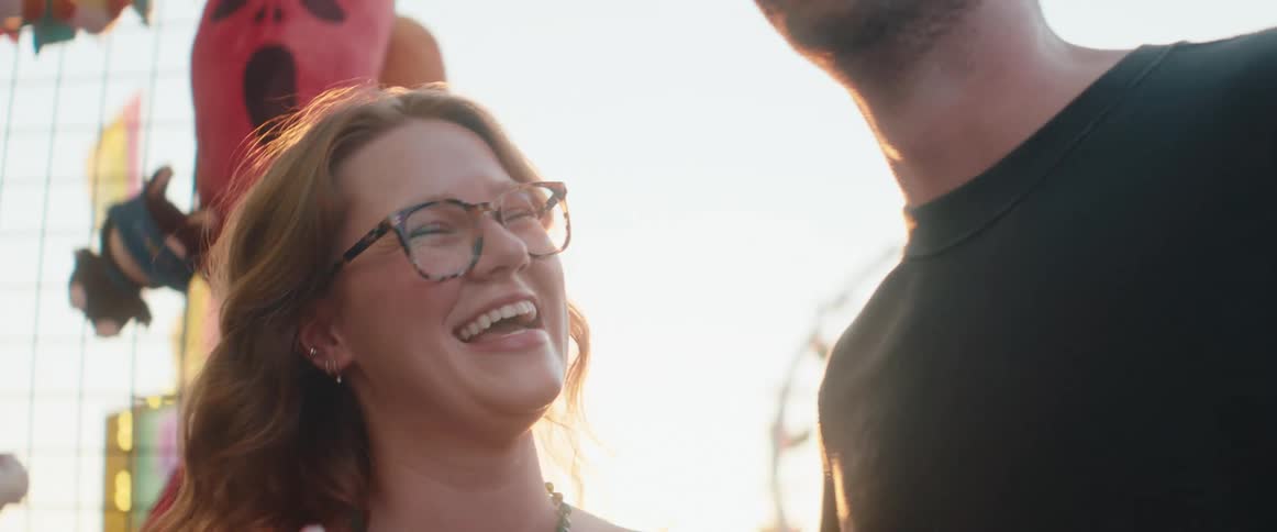 A mans hand gives a small pink stuffed animal to a woman wearing eyeglasses in the foreground and the woman and a man standing next to her both turn their heads to laugh loudly while backlit by bright daylight